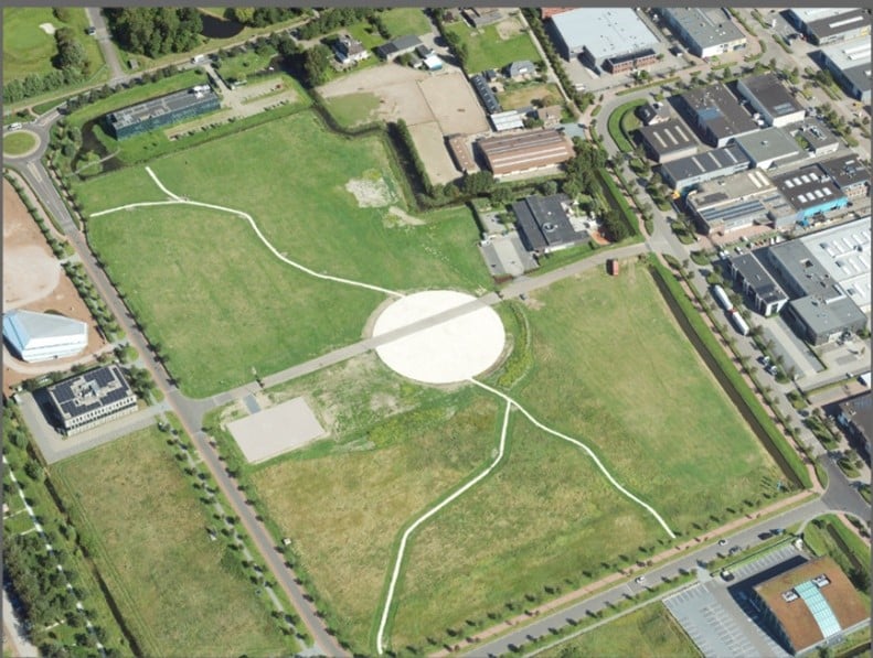 Luchtfoto van de campus met een groot open grasveld, een cirkelvormig plein in het midden en kronkelende paden, omringd door gebouwen en wegen.